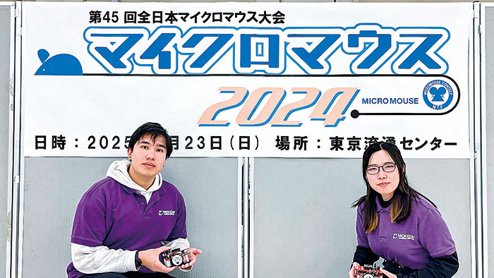 全日本マイクロマウス大会の看板前でマイクロマウスを手にする学生2名