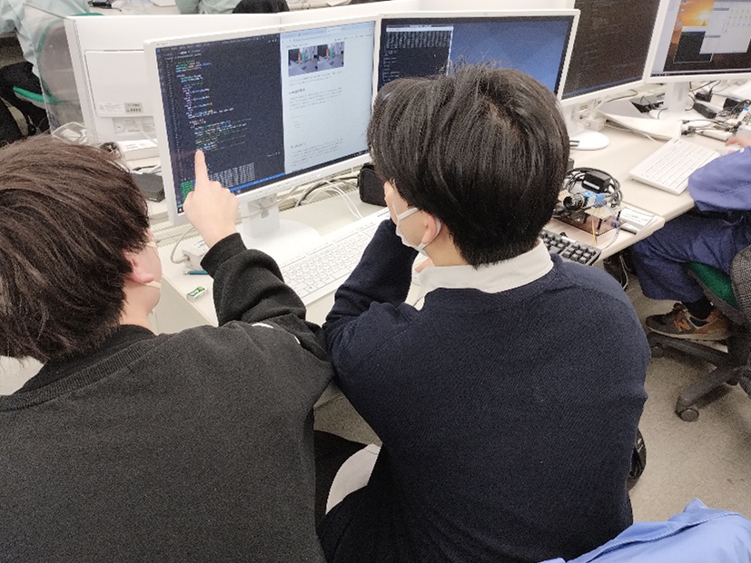 組込みソフトウェアプログラミングの授業風景。講師と生徒がPCでプログラミングしている。