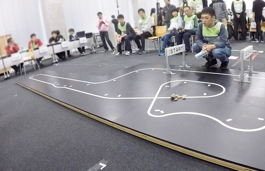 写真：競技会でコースを走行するマイクロマウスと制御する学生の様子
